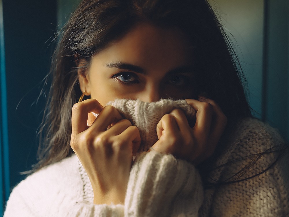 Vrouw trekt een crèmekleurige gebreide trui over haar kin in zacht raamlicht, een intieme portretfotosessie