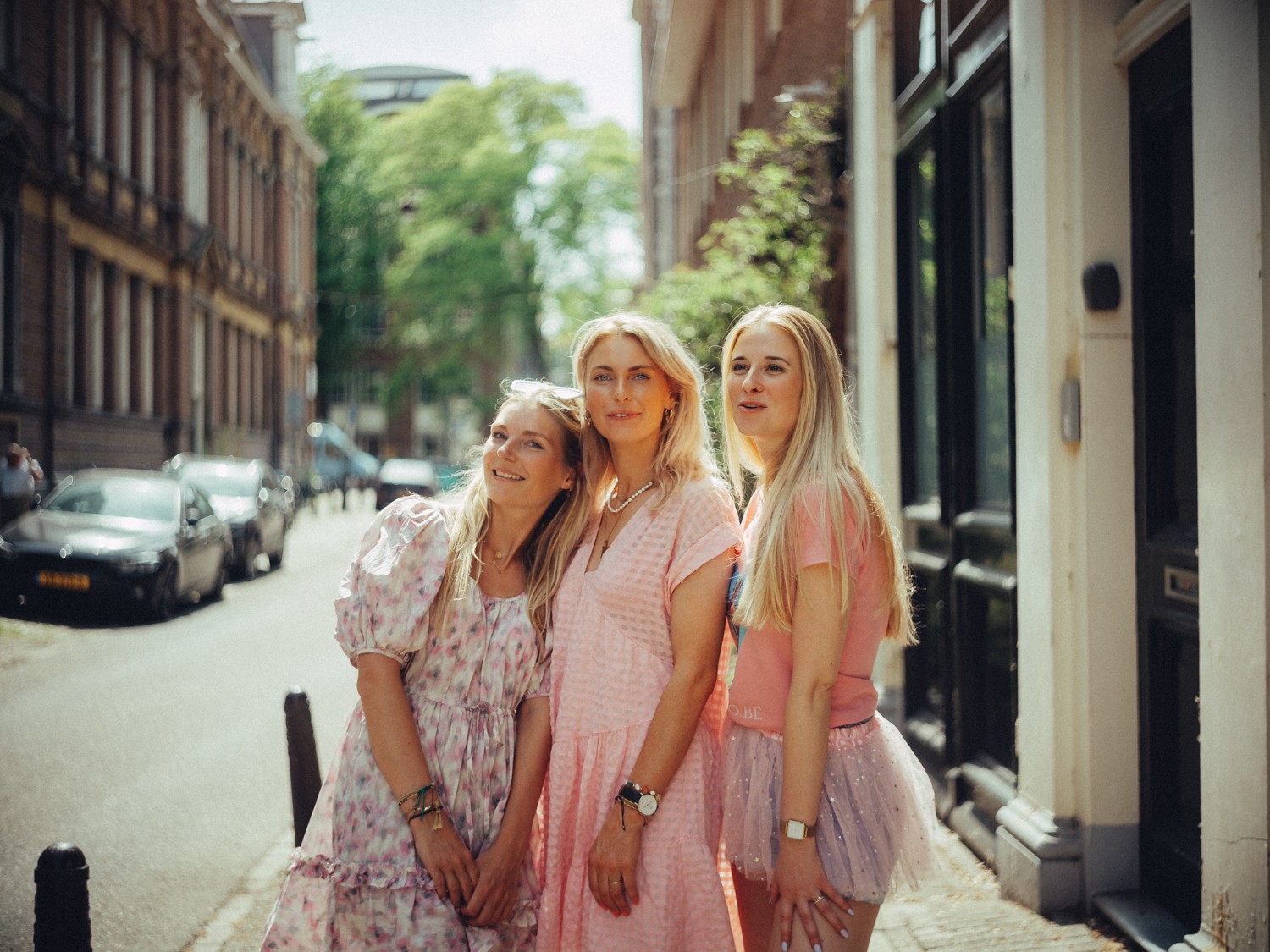 Three friends leaning together in the sun on a quiet street, in a pink dress, a floral dress and a pink tulle skirt