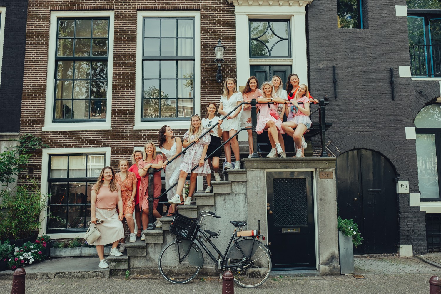 Bachelorette friends in pink and white on the stoop and railing of a brick canal house, a black bike parked by the door