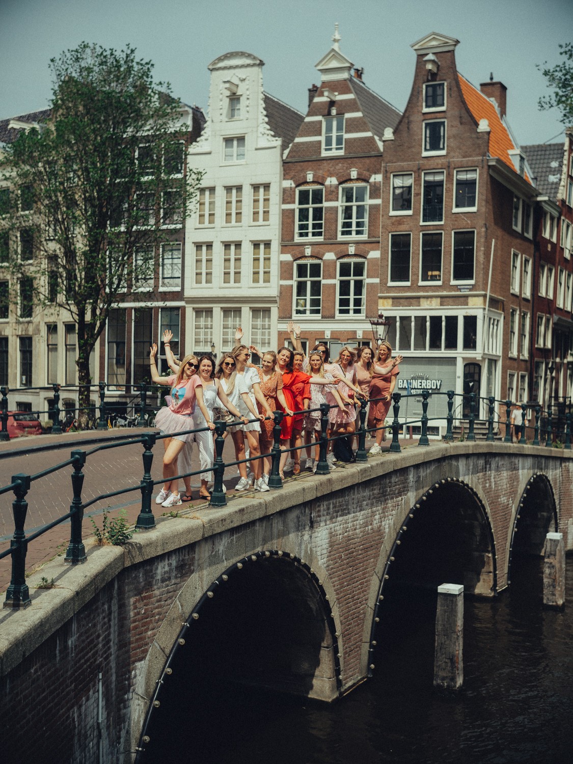 Bachelorette friends in pink and red waving from the railing of a three-arched brick bridge, gabled canal houses behind