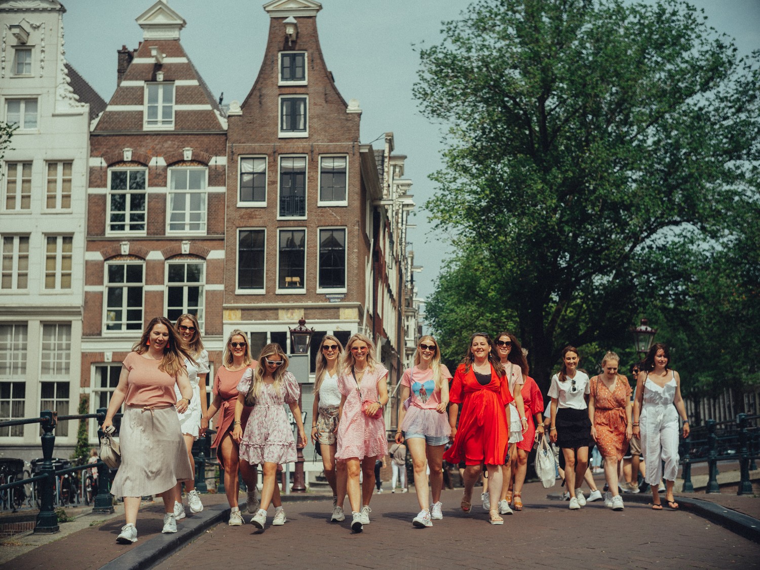 Group of friends walking toward the camera across a canal bridge, tall gabled houses and a big tree behind them