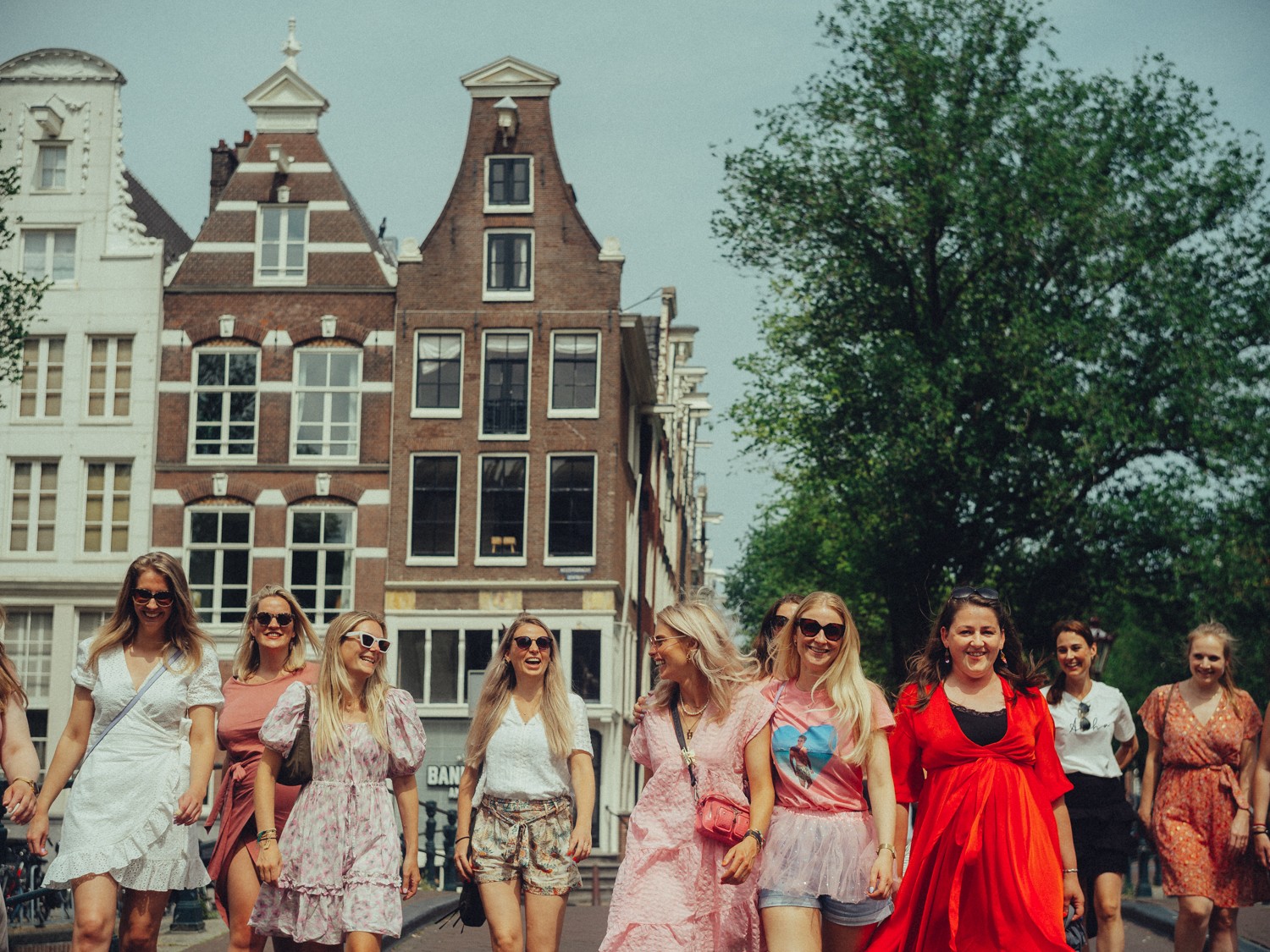 Friends in sunglasses laughing as they cross a bridge, a pink tulle skirt and a bright red dress in front of brick gables