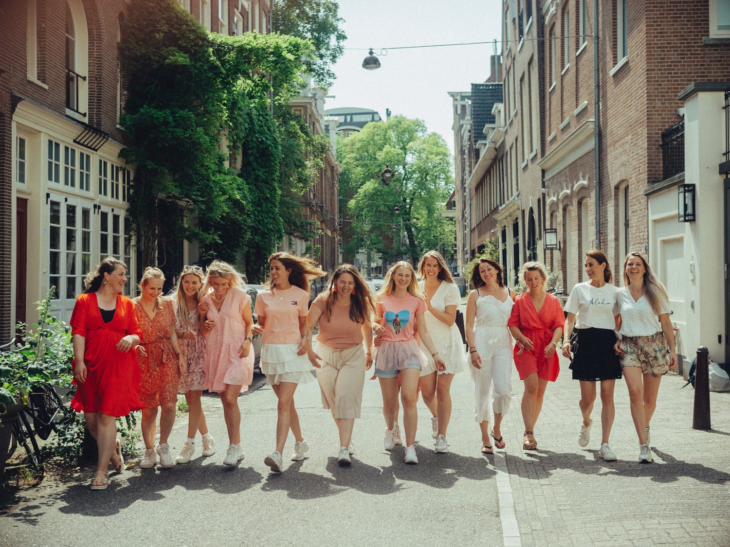 Twelve friends walking arm in arm down a sunny side street, hair flying, a red dress at one end of the line