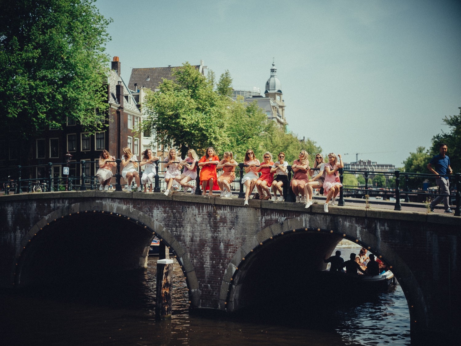 Row of friends seated on a bridge railing as a canal boat passes under the arch, a domed tower in the distance