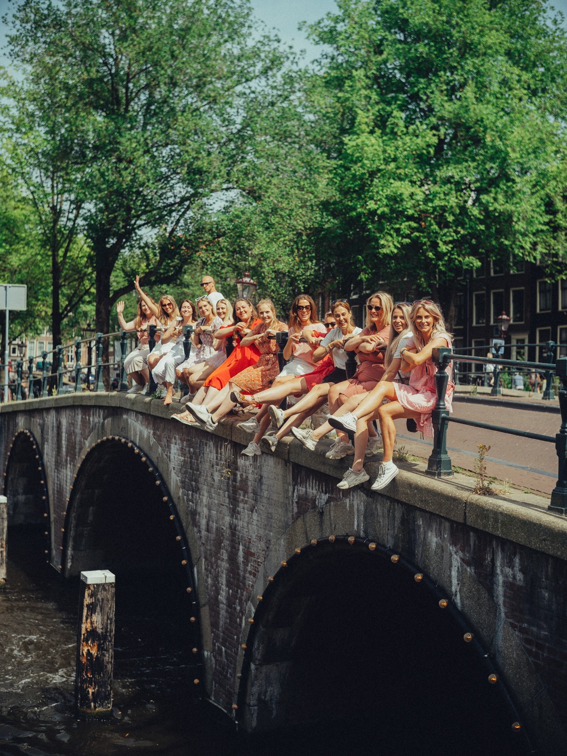 Friends sitting in a long row on the edge of an arched canal bridge, legs stretched out, under tall summer trees