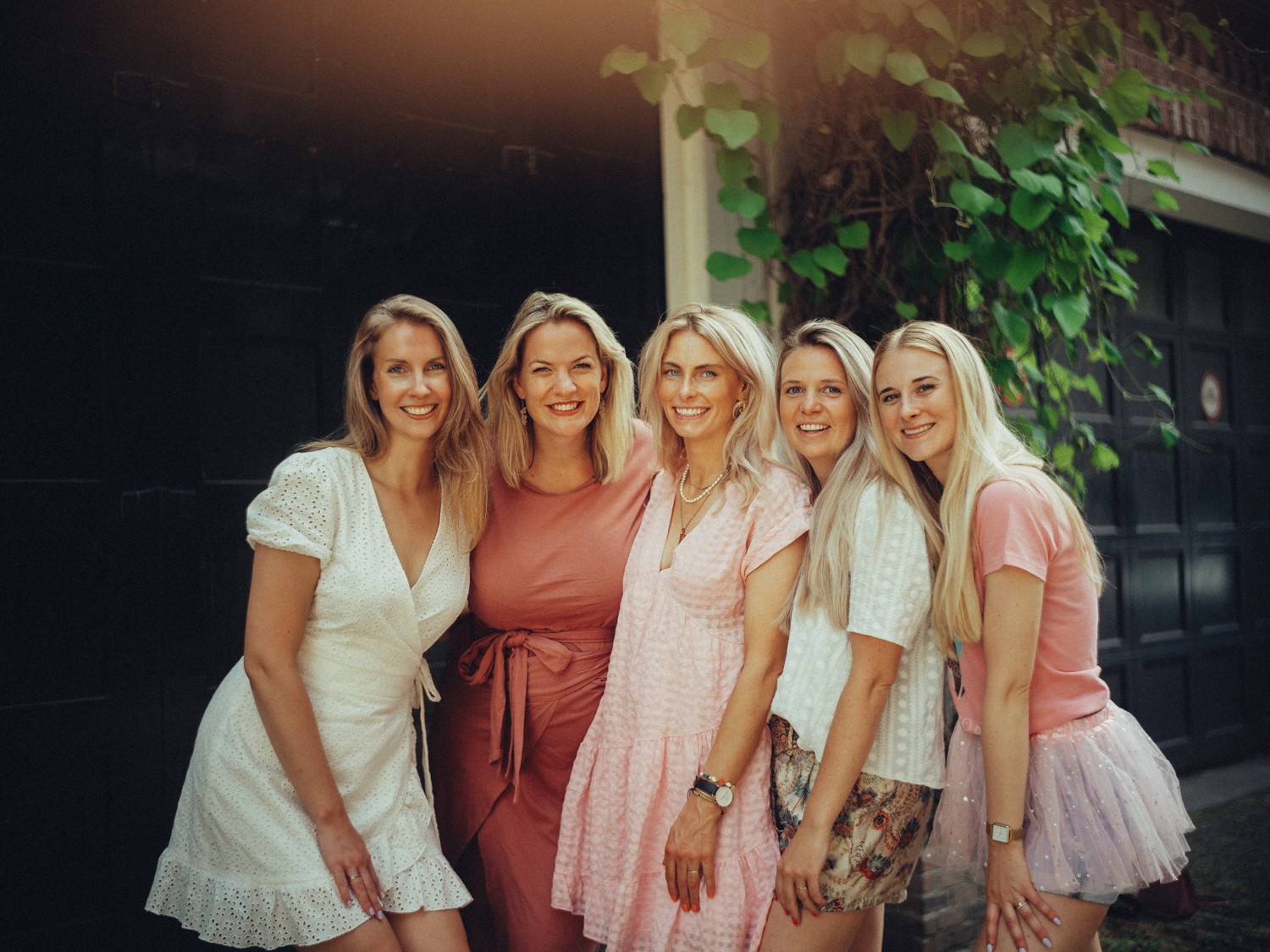 Five friends in white, coral and pink dresses standing close in front of a dark door, ivy and warm light above them