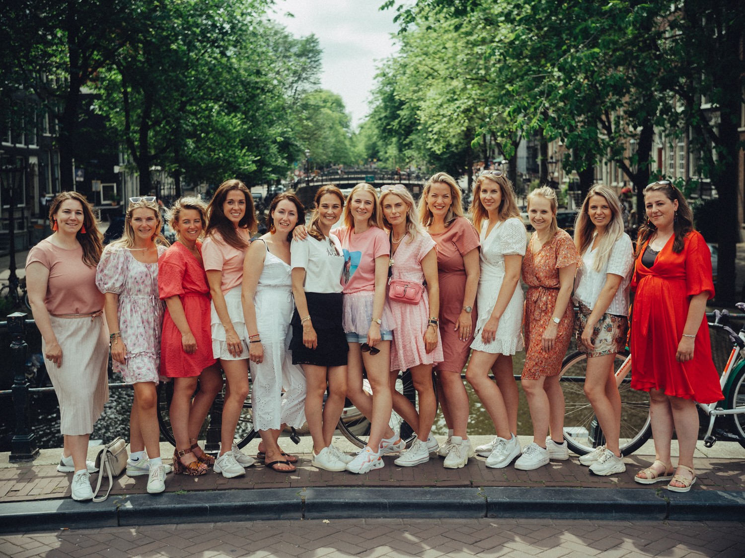 Thirteen friends at a bachelorette lined up on a canal bridge in pink, red and white summer dresses, tall trees over the water