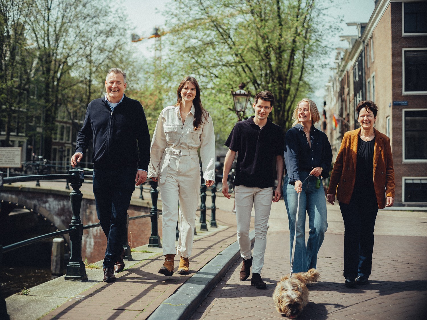 Gezin loopt langs de leuning van een brug naar de camera toe, met hun ruigharige terriër die vooruit draaft