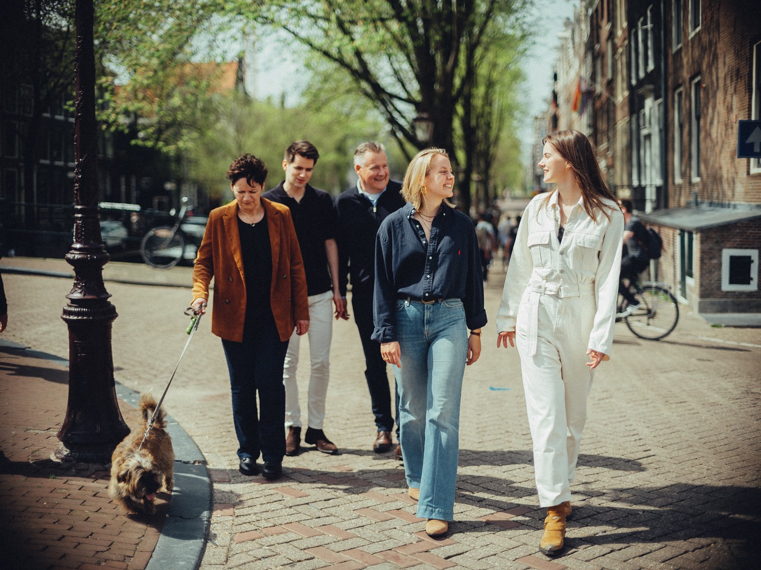 Gezin wandelt samen door een grachtenstraat in de lente, de moeder voorop met de terriër aan de lijn