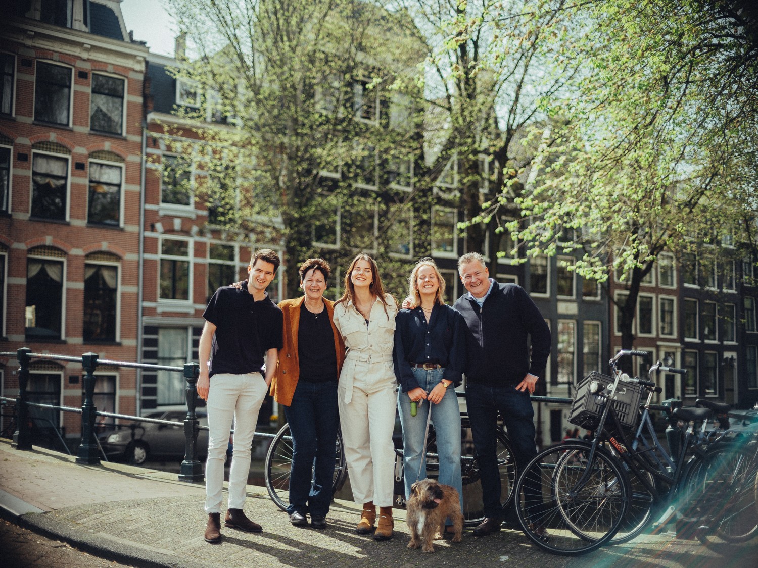 Gezin van vijf en hun terriër op een rij bij fietsen op een grachtenbrug in de lente, met bomen vol fris groen erachter