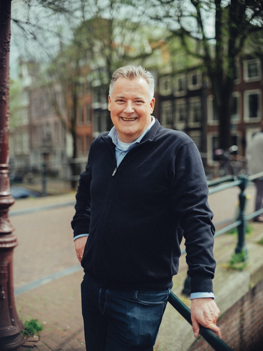 Vader in een marineblauwe trui met rits lacht bij de leuning van de gracht op een zonnige lentedag, met groene bomen erachter