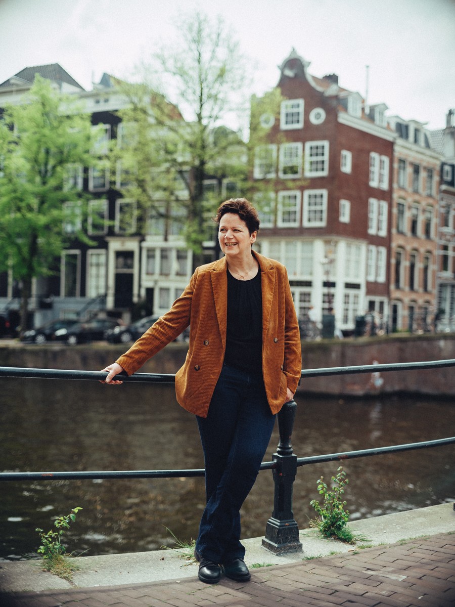 Moeder in een roestbruin suède jasje leunt ontspannen en lachend op de leuning van de gracht, met water en groen erachter