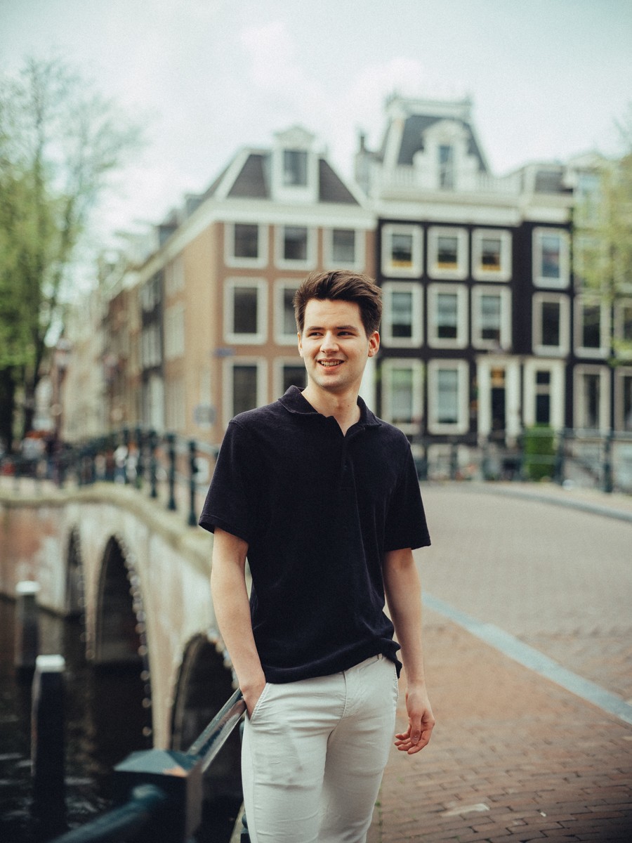 Zoon in een zwarte polo en witte broek staat alleen op een boogbrug over de gracht en lacht naar de camera