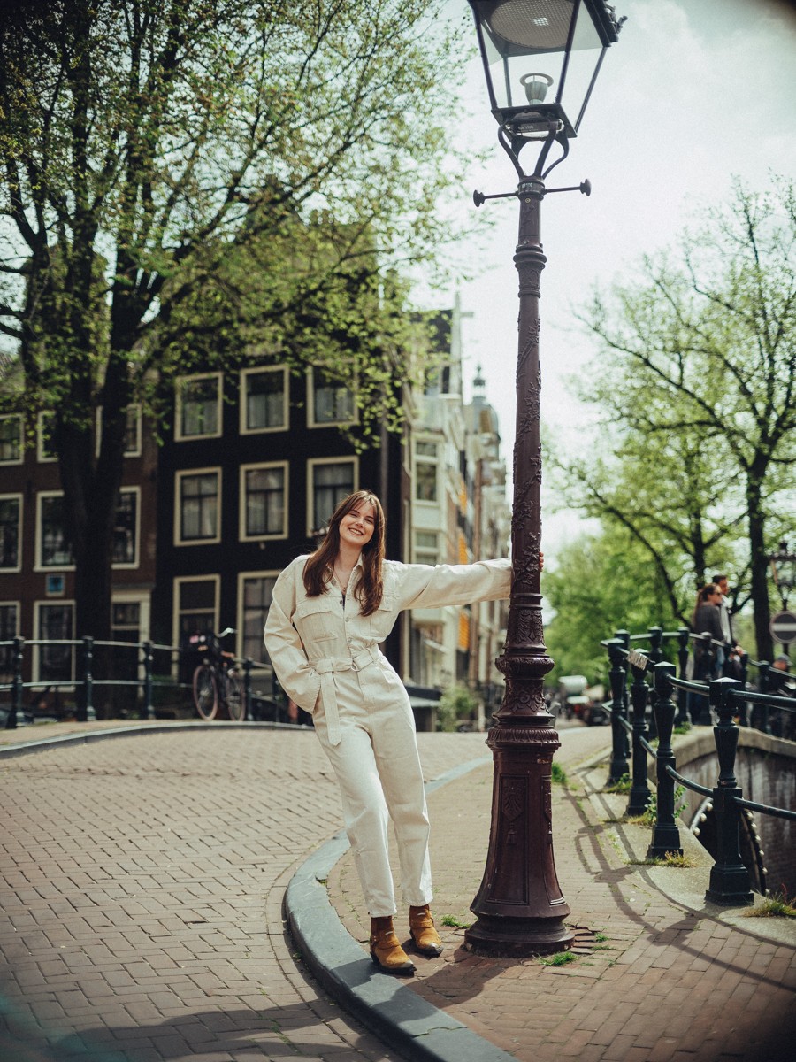 Dochter in een witte jumpsuit en bruine laarzen leunt tegen een lantaarnpaal langs de gracht in de lente