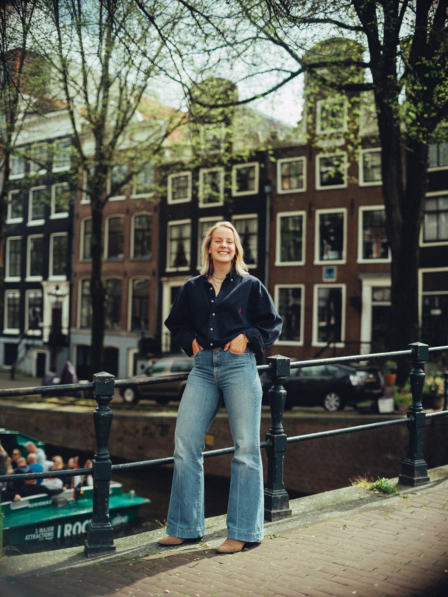 Dochter in een marineblauwe blouse en wijde jeans met haar handen in haar zakken terwijl er een rondvaartboot voorbijvaart