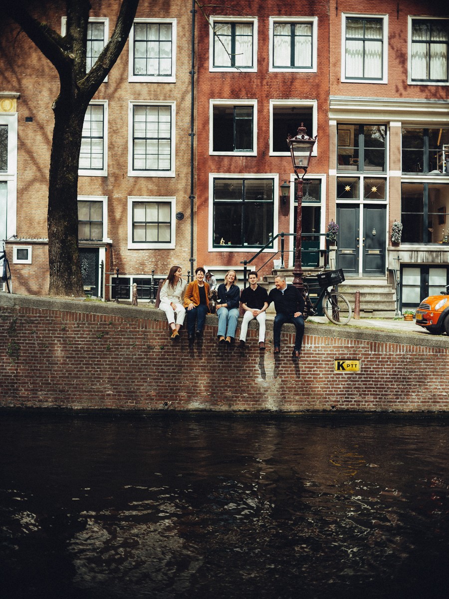 Gezin zit samen op de rand van een gracht, gezien vanaf de overkant, met bakstenen grachtenpanden erachter