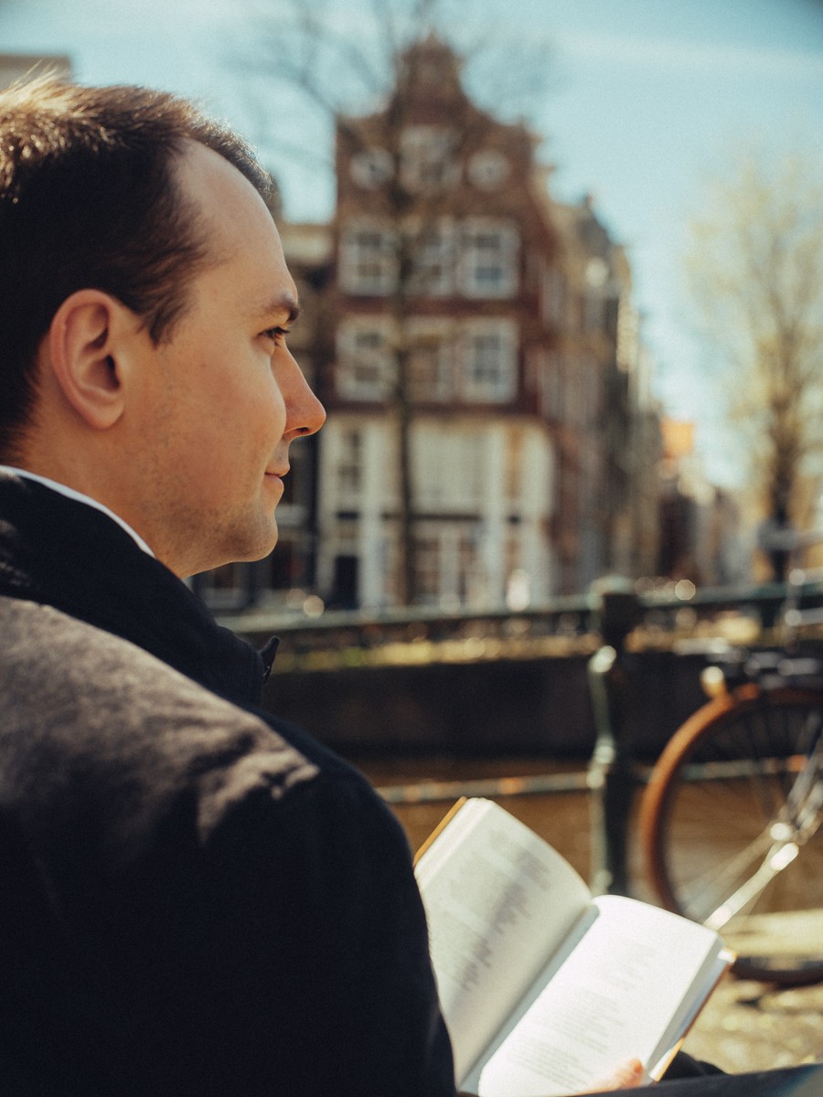 Man en profil leest een boek aan de gracht, met een rood bakstenen hoekhuis en kale bomen op de achtergrond