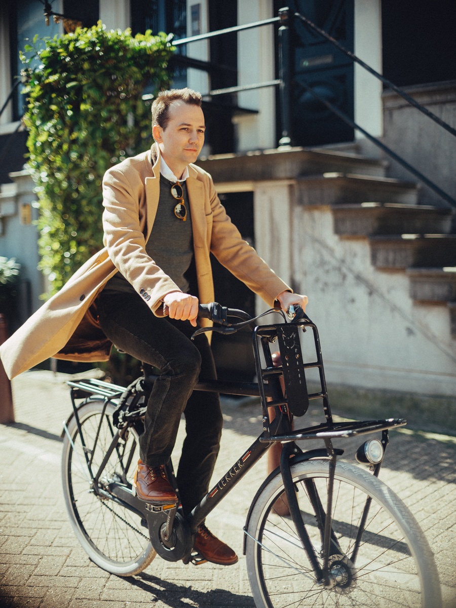 Man fietst langs witte stoeptreden en een haag, de camelkleurige jas open en bruine schoenen op de trappers