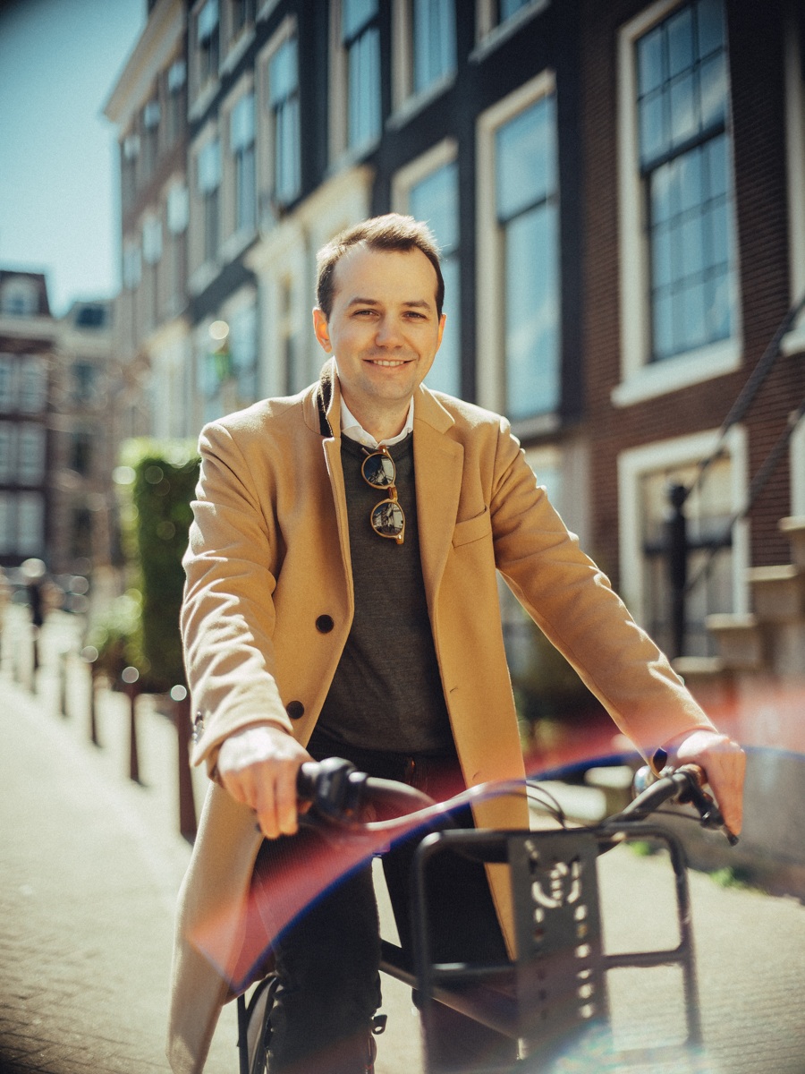 Lachende man in een camelkleurige jas met een zonnebril aan zijn trui, leunend op het stuur van zijn fiets