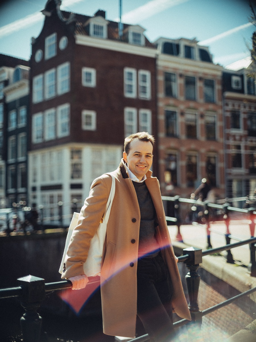 Lachende man in een camelkleurige jas met een canvas tas, leunend op de reling van een brug voor bakstenen huizen