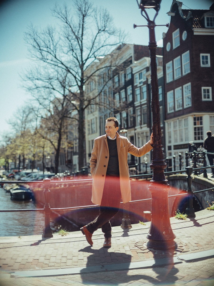 Man loopt langs een oude lantaarnpaal op een brug, de camelkleurige jas in beweging en een lichtvlek door het beeld