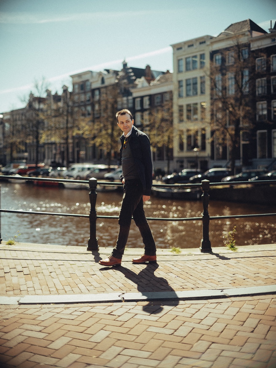 Man in een donkere jas loopt over een zonnige kade langs de gracht, zijn schaduw lang op de klinkers en bootjes op het water