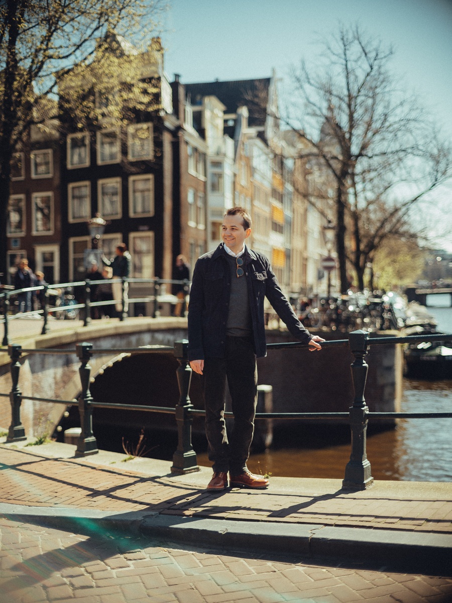 Man in een donkerblauwe jas op een brug over de gracht, een gebogen reling en huizen aan de overkant