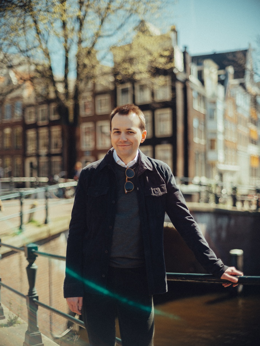 Man in een donkerblauwe jas bij de reling van de gracht, met zonnige trapgevels en kale bomen erachter