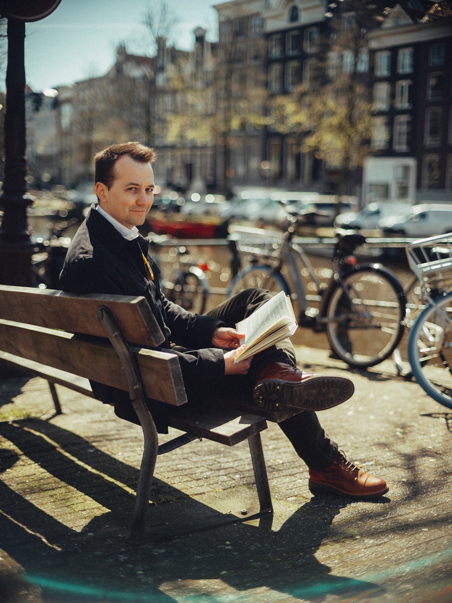 Man leest een boek op een houten bank, één been over het andere, fietsen en huizen aan de gracht onscherp erachter