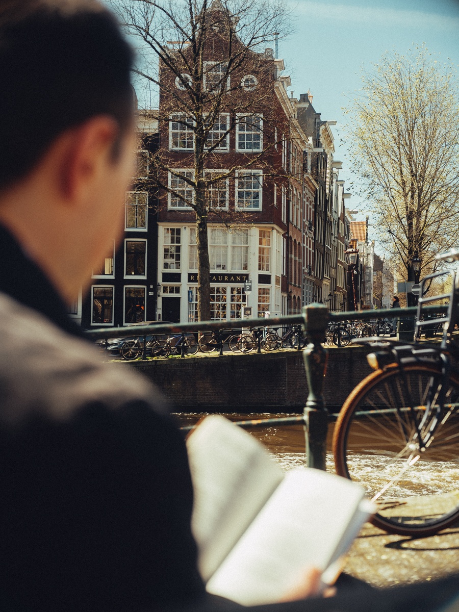 Blik over de schouder van een man die een boek leest bij de reling van de gracht, met een fiets en gevels verderop