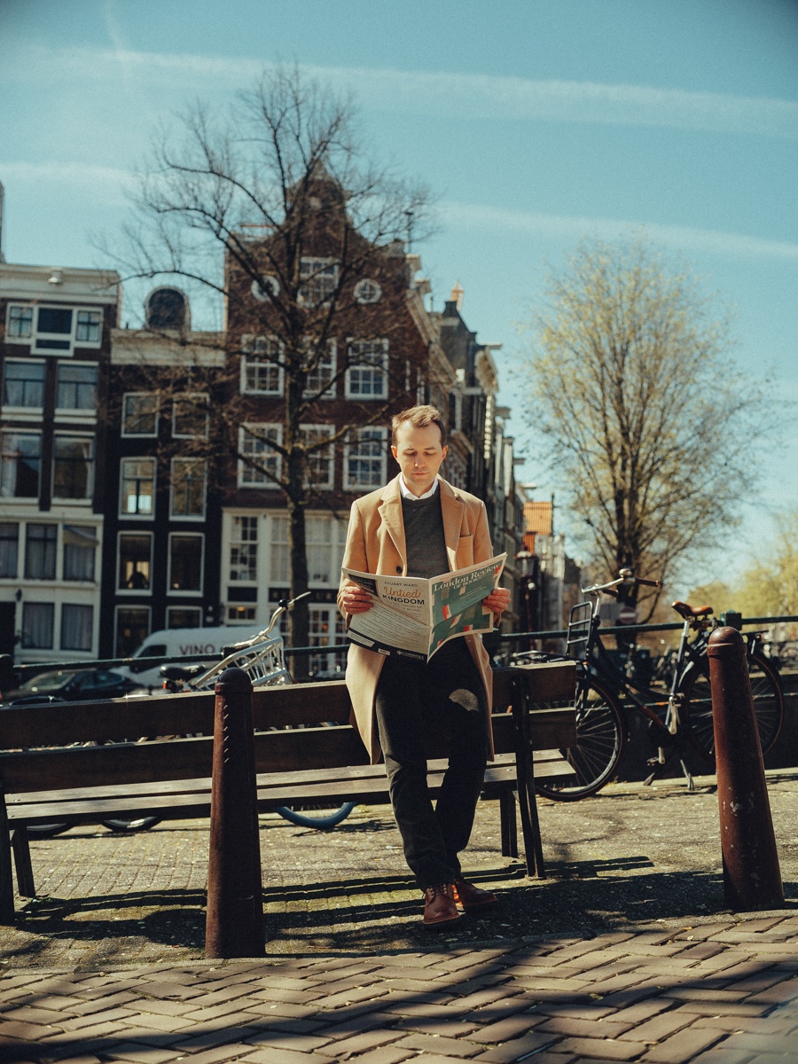 Man leest een krant op een bankje aan de gracht, tussen paaltjes, fietsen en hoge trapgevels