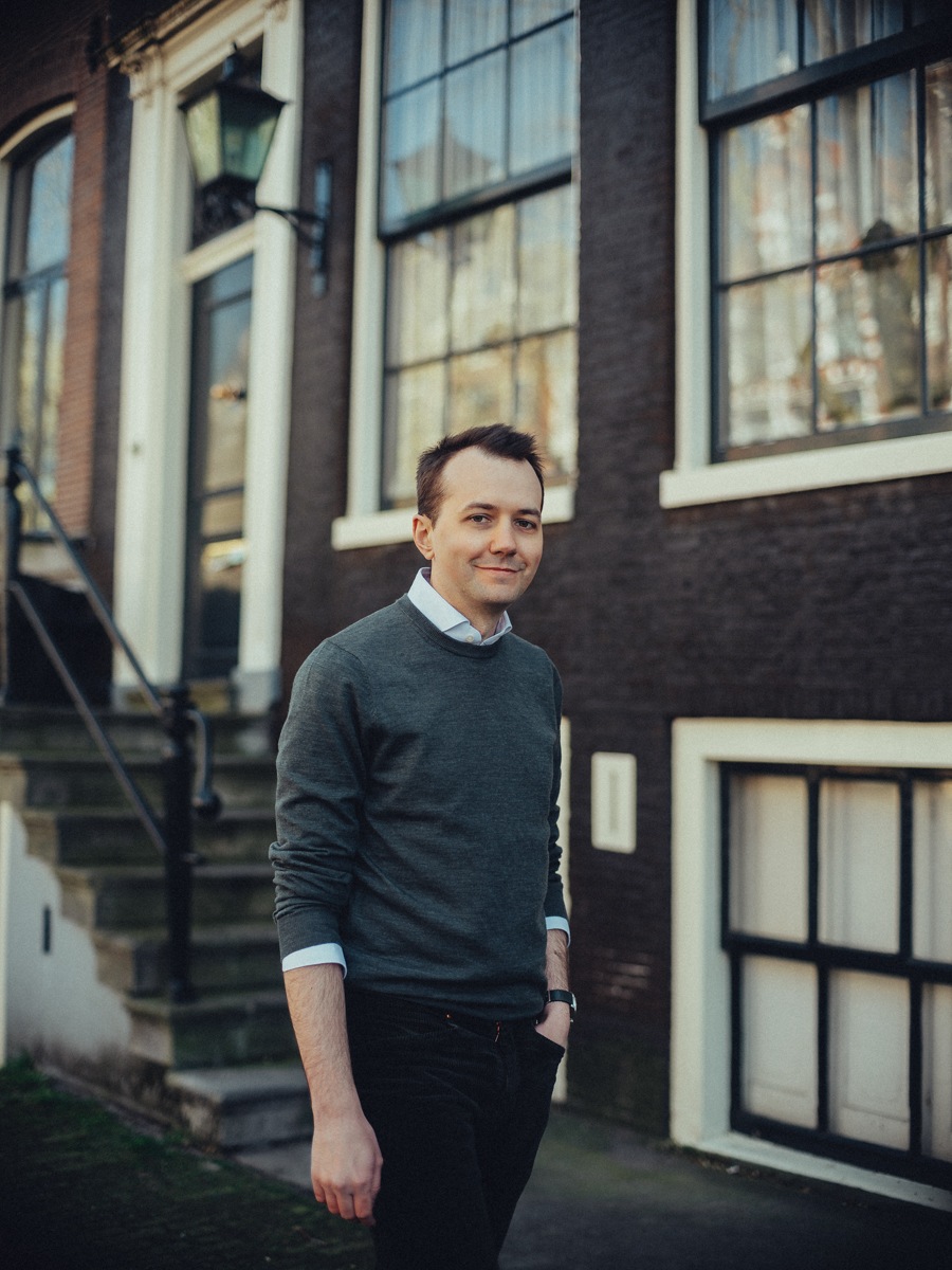 Man in een grijze trui staat bij de stoep van een bakstenen huis, de handen ontspannen langs zijn lichaam