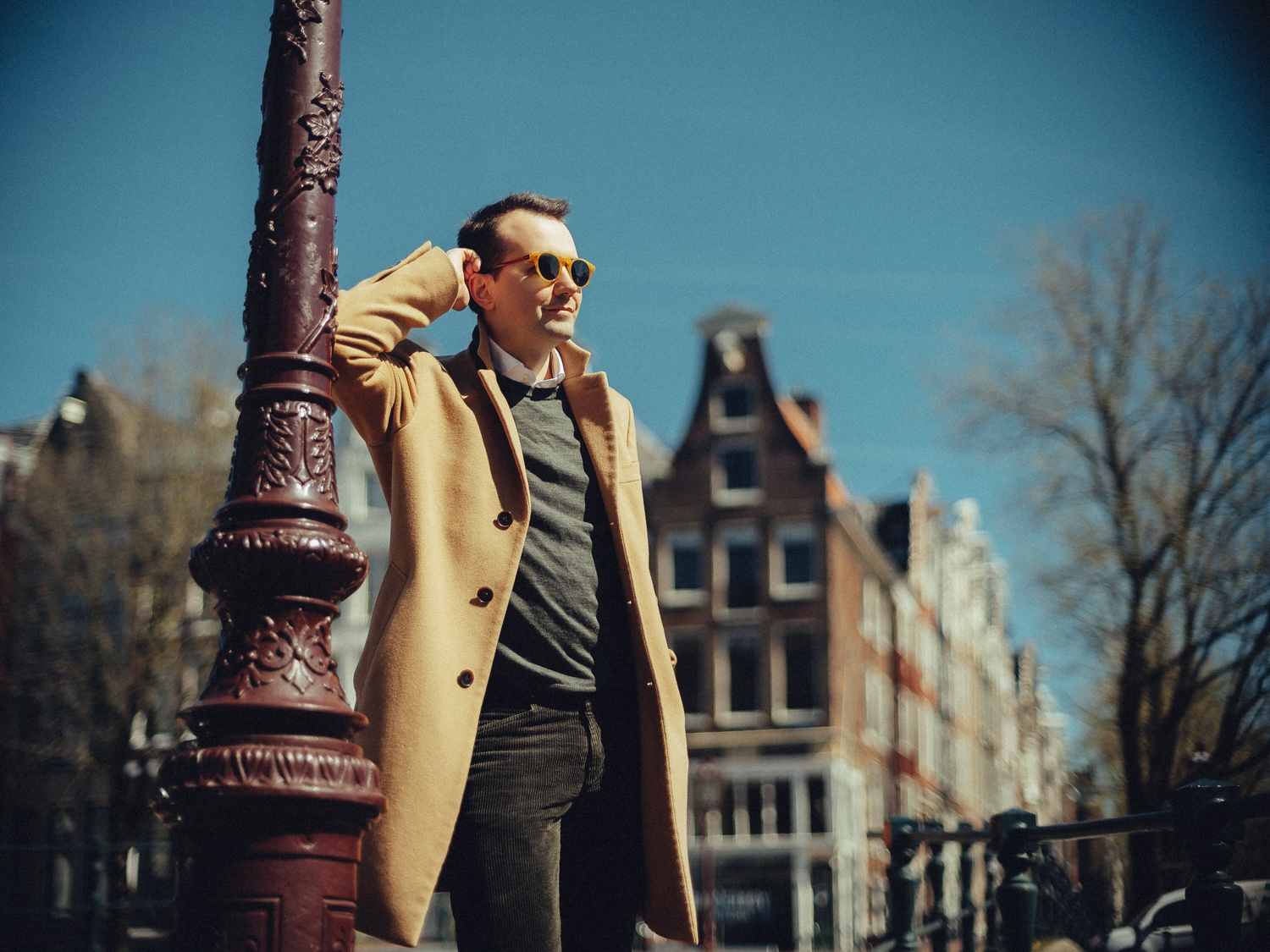 Man met zonnebril en camelkleurige jas leunt tegen een sierlijke lantaarnpaal, een hand bij zijn hoofd, met gevels erachter