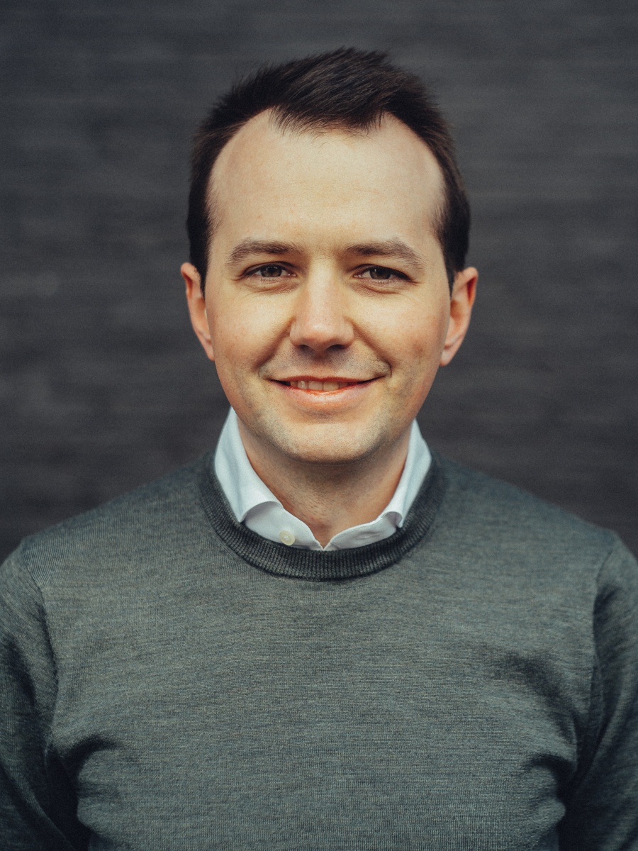 Portret van een lachende man in een grijze trui met witte kraag tegen een donkere bakstenen muur