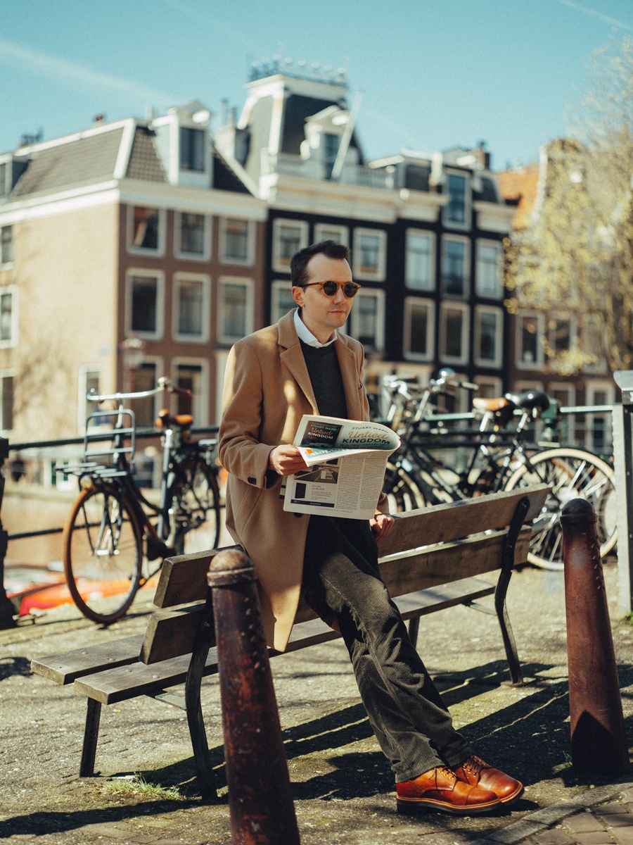 Man met zonnebril en camelkleurige jas leest de krant op een bankje, met fietsen en gevels in de zon
