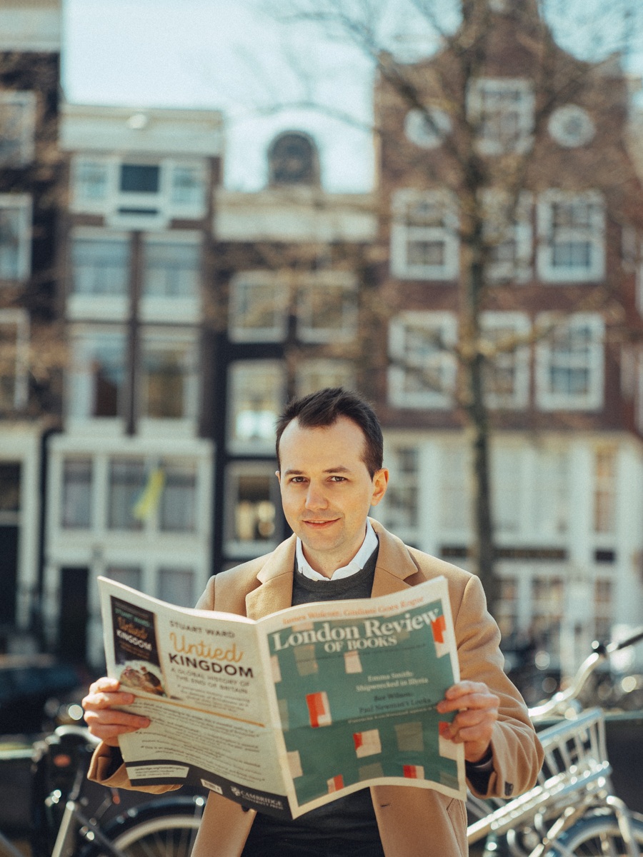 Man houdt de London Review of Books open naar de camera en lacht erboven uit, met grachtenpanden erachter