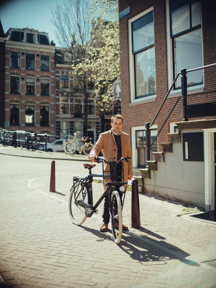Man staat met zijn fiets naast een stoep met paaltjes in een zonnige straat, een bloeiende boom op de achtergrond