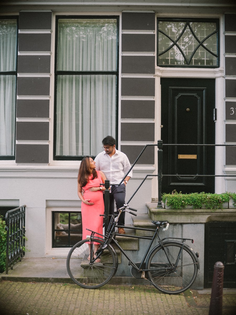 Stel bij de deur van een grachtenpand met een zwarte fiets tegen het hek, de koraalrode jurk fel tegen de grijze gevel