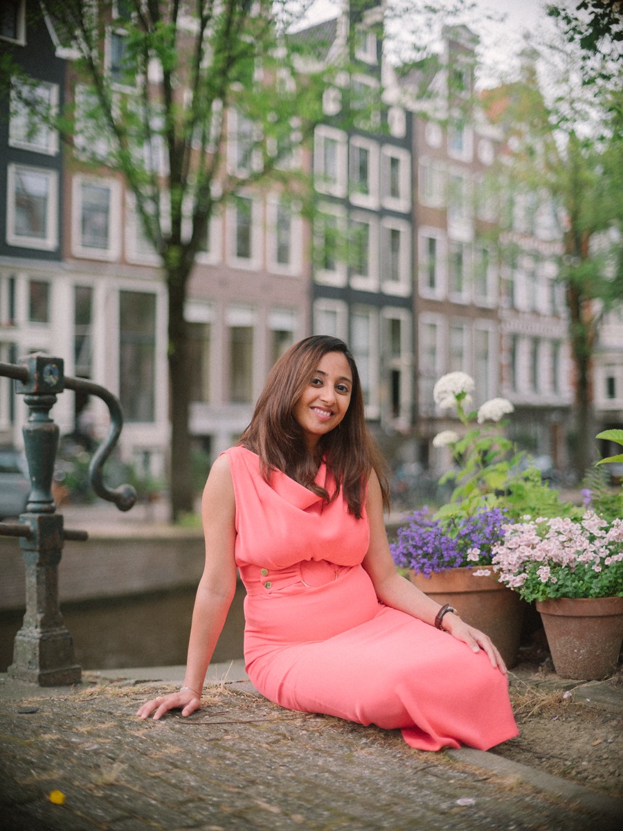 Zwangere vrouw zit bij bloempotten naast een gracht in de zomer, haar koraalrode jurk uitgespreid over de stenen