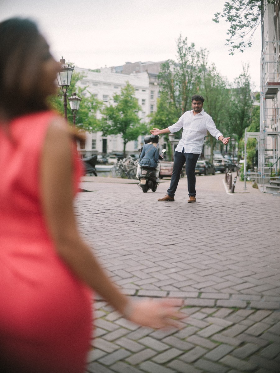 Vrouw in een koraalrode jurk loopt over een straat met klinkers terwijl haar partner in de verte zijn armen spreidt