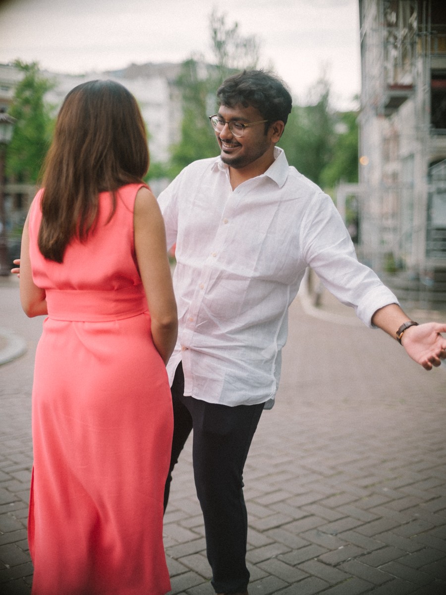 Lachend stel in een straat met klinkers, hij gebaart speels terwijl zij in haar koraalrode jurk naast hem loopt
