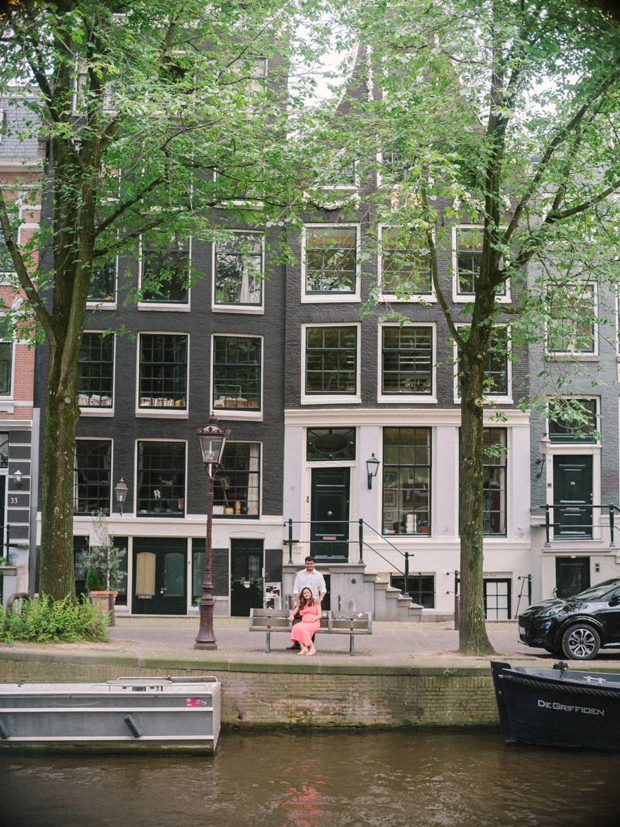 Stel klein in beeld op de stoep van een grachtenpand, met een gracht vol bomen en een aangemeerd bootje ervoor