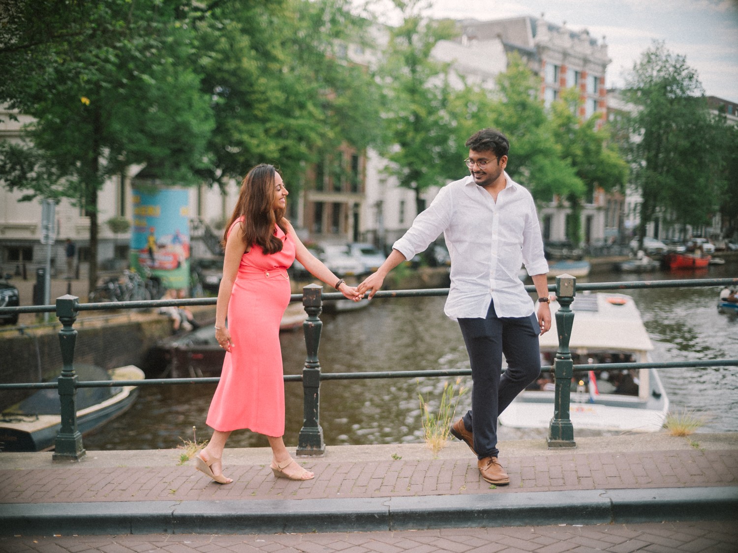 Stel loopt hand in hand over een grachtenbrug, een rondvaartboot eronder en bomen vol blad in de zomer