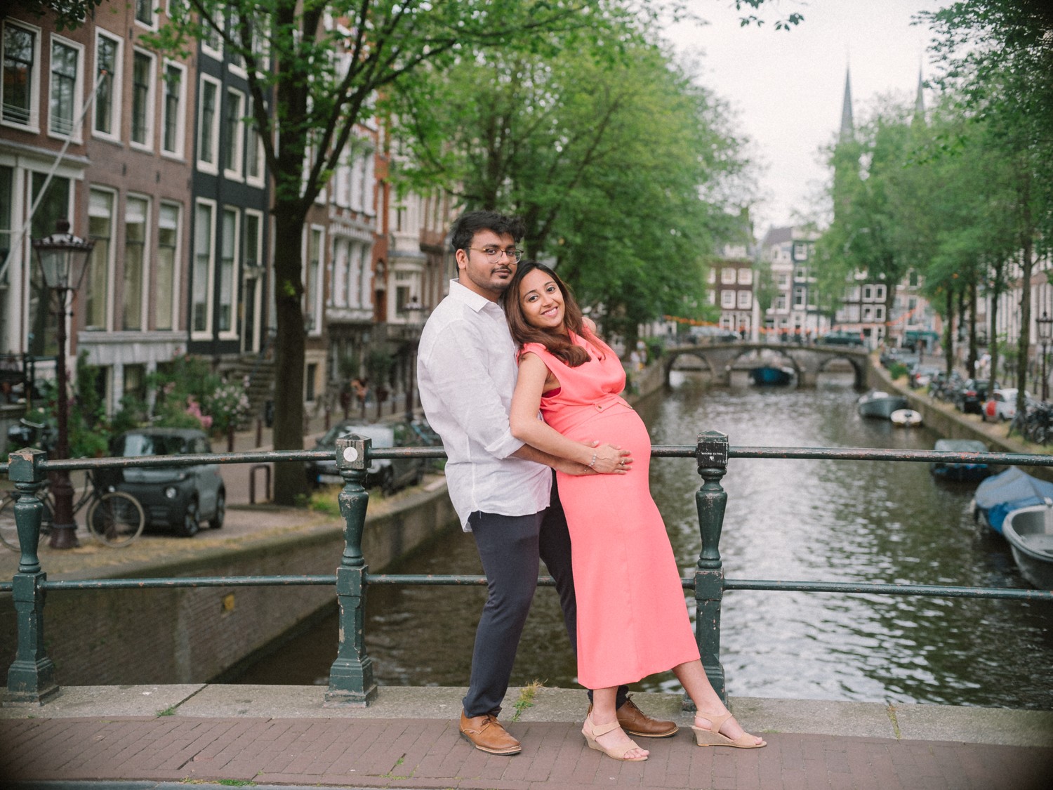 Stel omhelst elkaar op een grachtenbrug, zijn handen op haar buik, de bomen vol blad in de zomer langs het water