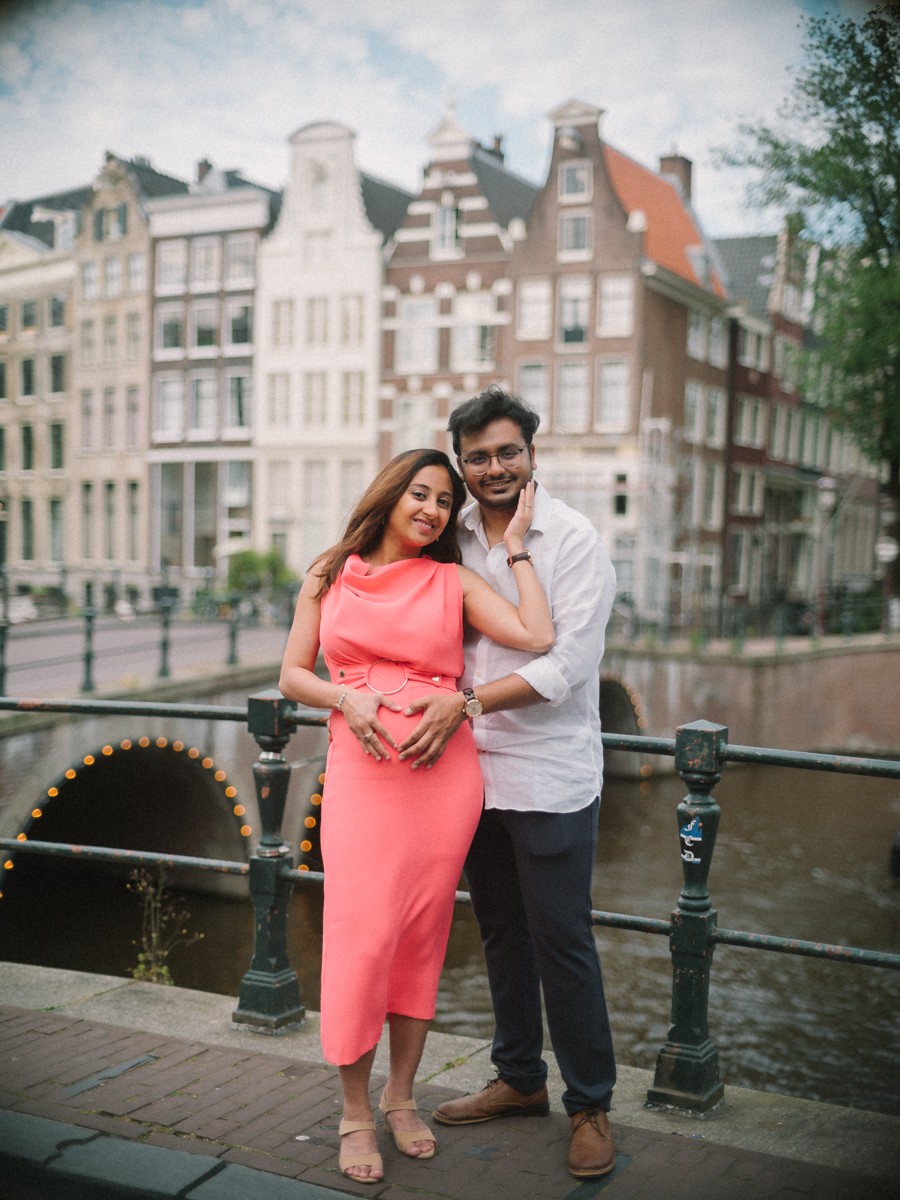 Stel op een grachtenbrug met zwarte hekken, een rij gevels erachter tijdens een zwangerschapsshoot in de zomer
