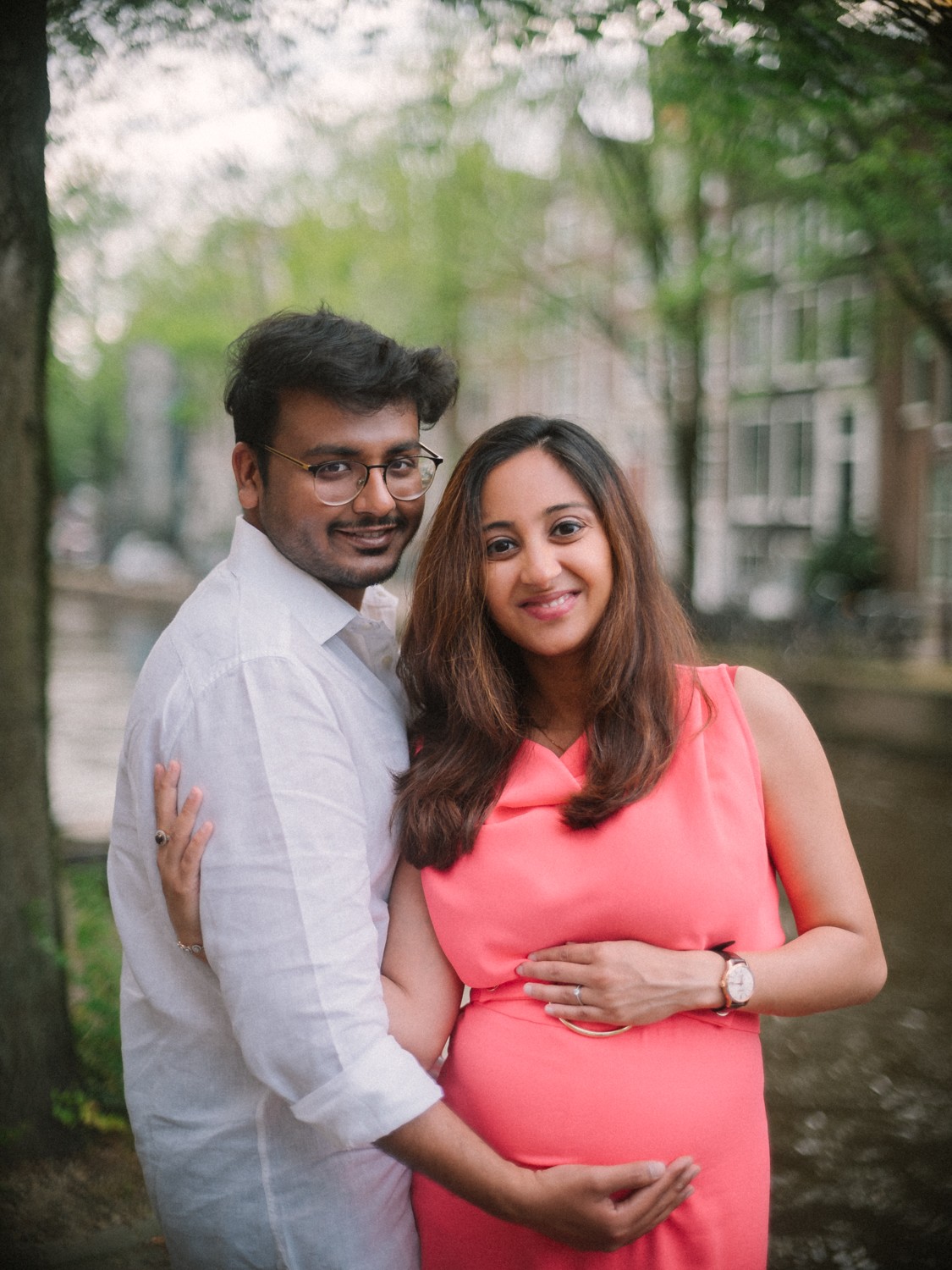 Stel lacht naar de camera, zijn hand op haar buik en die van haar erbovenop, groene bomen aan de gracht in de zomer