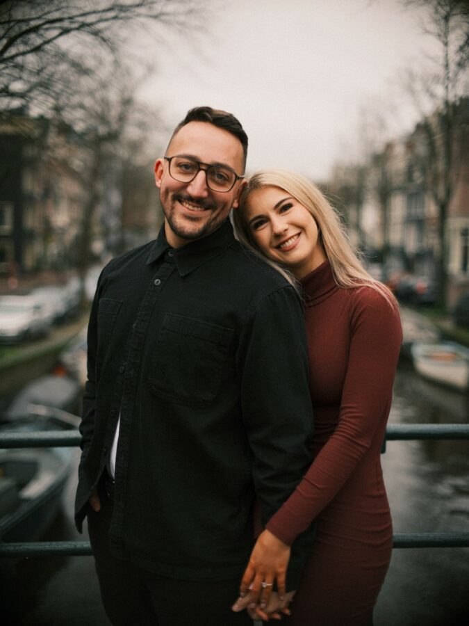 A smiling couple holding hands on a canal bridge showing off a new ring during their Amsterdam engagement shoot in winter