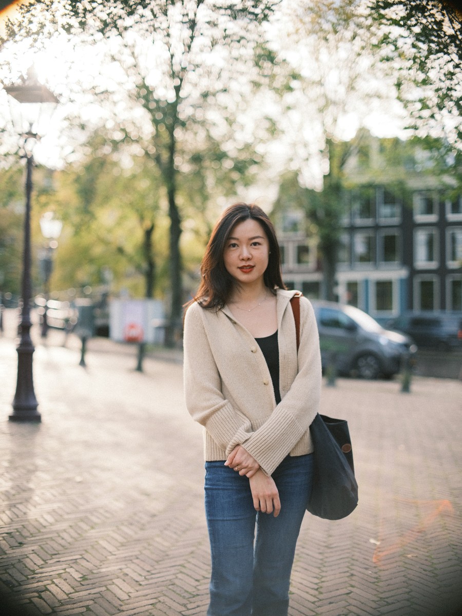 Portret tot de heupen van een lachende vrouw met rode lippen en gevouwen handen, een beige vest over een zwarte top