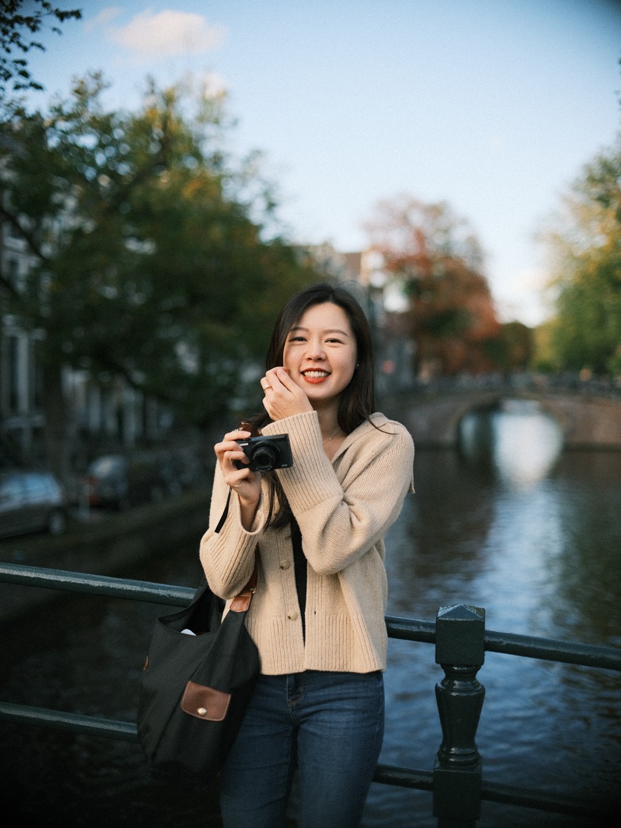 Vrouw lacht op een grachtenbrug met een kleine zwarte camera in haar handen, een hand tegen haar wang en het water erachter