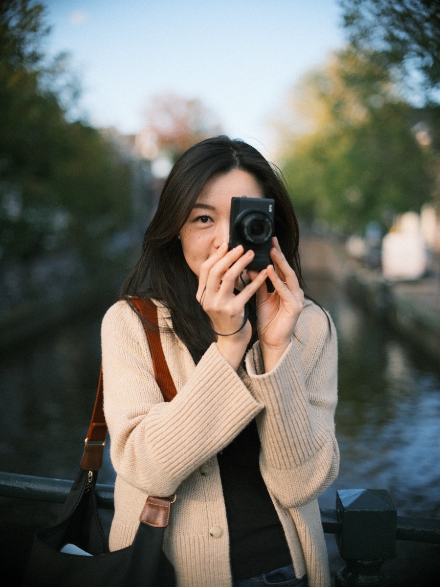 Vrouw richt haar compacte camera recht op de lens, met één oog ernaast, herfstbomen en een gracht wazig op de achtergrond